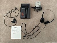 Photo showing vintage Sony Walkman cassette player, single earbud, headset with microphone, and power adapter on carpet.