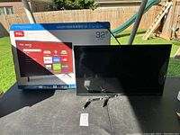 32-inch TCL Roku Smart TV on table with original box behind it outdoors. TV screen is off and two detachable stand legs are placed in front.