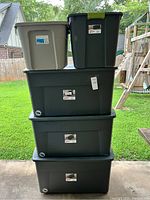 Five plastic storage bins stacked outdoors, four large black Sterilite bins and one smaller beige bin on top