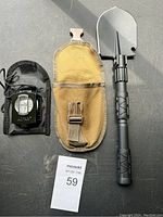 Photo shows the high quality compass in a black nylon case with a belt loop and measurement dial, alongside the black collapsible shovel partially assembled with its tan-yellow fabric carrying pouch visible.