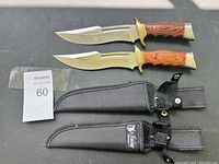 Two collectible cutting knives with wood handles and brass hand guards placed above two black nylon sheaths with button snaps.