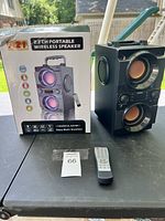 Photo showing the A21 2.2 CH portable wireless speaker, front view, placed next to box and remote control on a table outdoors.