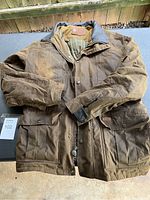 Full view of the brown oilskin coat laid flat showing front pockets, zipper and snap-button closure, collar, and sleeves.