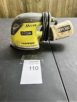 Front angle view of Ryobi orbital sander showing main body, grip, and attached dust bag. Cord wrapped around sander body.