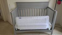 Gray child's day bed with white mesh safety rail attached, situated indoors against a wall and carpeted floor.