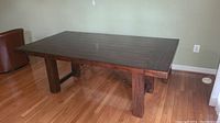 Front angled view of wooden dining table showing broad surface and thick legs, set on hardwood floor.