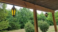 Two amber glass and metal hanging lanterns mounted on outdoor wooden beam against green trees background