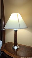 Front view of one table lamp lit, showing white fabric shade, wooden and brushed metal base