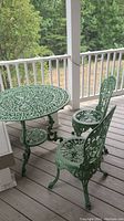 Full view of green painted metal outdoor round table and two matching chairs on a wooden deck, showing intricate openwork designs.