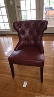Front view of the merlot-colored tufted accent chair showing button tufting and nailhead trim on edges, wooden legs visible, placed on hardwood floor near glass doors.