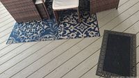 Blue polyurethane outdoor rug with white floral and geometric patterns, furniture on top, visible wear and small hole.