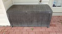 Front view of large brown plastic outdoor storage bin with woven wicker texture, sitting on a brick patio.