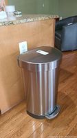 Side view of the Simplehuman metal kitchen trash can next to counter and hardwood floor, showing the full height and foot pedal.