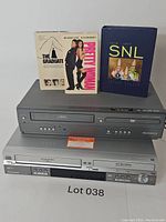 Magnavox and Panasonic DVD/VCR combo players stacked with Pretty Woman VHS, The Graduate VHS, and SNL Second Season DVD box set on top.