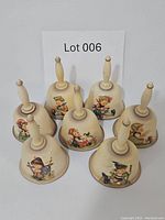 Seven Goebel Hummel porcelain collectible bells with cream-colored bases and painted children characters, shown with Lot 006 sign