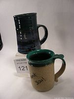 Photo showing two pottery mugs side by side, one dark greenish-blue mug with a glossy glaze and the other textured beige mug with green glaze on rim and handle and 'Thank you!' writing.