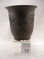 Front angle of a black stoneware bonsai pot with carved floral and geometric patterns, multiple drainage holes visible, on a stand with auction tag.