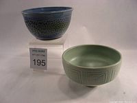 Two pottery bowls side by side; the green matte bowl with decorative pattern and the blue textured bowl on a stand with business card.
