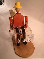 Front view of the wooden jigger puppet seated on a birch paddle, showing the figure painted in bright colors with a yellow hat and the paddle base.
