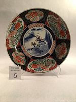 Front view of large round porcelain Imari charger with intricate hand painted panels of floral designs and central cobalt blue landscape with floral motifs.