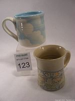 Two pottery mugs, one with blue sky and white cloud design (Patti Willis) and another with animal/star pattern (Beardsley) on white stoneware, standing next to each other.