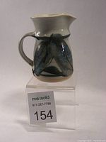 Front angled view of Robin Hopper cream jug with blue and green leaf-like motif on a white base, showing handle and spout.