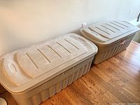 Two beige Rubbermaid Roughneck jumbo storage totes with lids, placed side-by-side on wooden floor, showing size and shape.