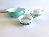 Three vintage mixing bowls arranged on white surface. Front left is small white Hazel Atlas bowl with green ivy leaf pattern on exterior. Center right is clear white Pyrex Amish Butterprint pattern bowl with tab handles. Rear left is large Robins Egg blue Pyrex bowl with tab handles.