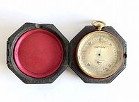 Open antique aneroid barometer housed in a hexagonal hinged case showing the barometer face with markings and needle, interior case lining red fabric