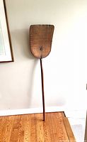 Full view of antique wooden maple syrup paddle leaned against wall, showing entire length and scoop shape.