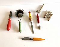 All seven vintage kitchen utensils in one image including peeler, serrated knife, melon baller, strainer, honing tool, and clothespins spread out on white surface.