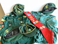 Lot overview showing two green Boy Scouts shirts with patches, two green wolf cub hats with yellow piping, two green berets, and red merit banner.