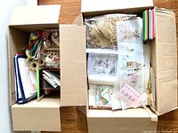 Top-down view of two open cardboard boxes containing assorted craft and office supplies packed inside.