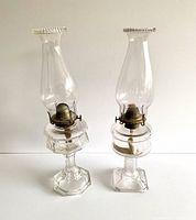 Full view of two vintage pressed glass coal oil lamps side by side, both with chimneys and brass burner fittings