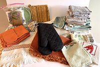 Overview of assorted kitchen linens including placemats, napkins, tea towels, hot pads and oven mitts arranged on a table.