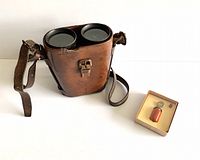 Binoculars in hard leather case with shoulder strap and boxed bird call placed beside them