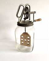 Front view of full butter churn showing glass jar, wooden paddle inside, metal lid with gear mechanism and wooden handle.