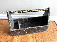 Front side view of the vintage wooden tool tote showing heavily chipped layers of black, green, and mustard paint, rectangular shape, and handle.