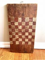Front side of thick wooden rectangular board showing hand-painted checkerboard pattern in faded off-white and rusty red squares. Multiple signs of wear and age, scratches, dents, and rustic surface.