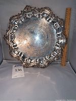 Front view of ornate silver tray showing detailed Baroque design and engraved center with ruler beside for scale.