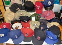Photo showing assortment of vintage and newer baseball caps including sports teams like Boston Red Sox, Philadelphia Phillies, San Francisco Giants, and costume hats like police hat and bowler hat.