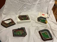 Six miniature baseball stadium replicas displayed on white cloth including Fenway Park, Ebbets Field, and Wrigley Field models in various packaging and conditions.