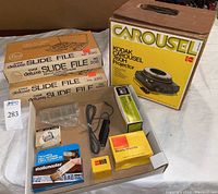 Photo showing Kodak Carousel 760H projector box, two Logan metal deluxe slide file boxes, and a tray with various small items including slide mounts, cables, and cleaning tools.