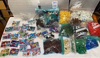 Wide view showing many bags of loose LEGO bricks sorted by color and multiple sealed mini LEGO sets in original packaging spread on table.