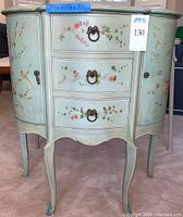Front view of the green hand-painted wooden nightstand showing floral and bird designs, three drawers with metal pull rings, and two curved cabinet doors with round knobs.