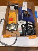 Box containing three digital cameras and Kodak lens cleaning papers with brand tags for Samsung, Kodak, Canon