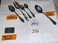 Photo showing four sterling silver spoons and one silver plated spoon arranged on white background with tags indicating '42g sterling' and 'silver plate', along with a small rectangular silver bar in a plastic cover tagged as 'Troy oz silver'.
