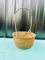 Front view of an antique cast iron smelting pot with a large metal handle. The pot shows signs of rust and wear consistent with its age and use in blacksmithing or metalwork.