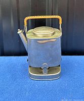 Front view of antique metal kettle with hinged lid, spout, and woven handle on blue surface.