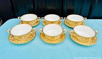 Overview photo showing six double handled cups and six matching saucers with gold ornate pattern on blue backdrop.